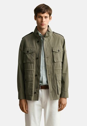 Junger Mann trägt eine olivgrüne Jacke mit mehreren aufgesetzten Taschen vorne, ein gestreiftes Hemd, weiße Hose und einen braunen Gürtel.