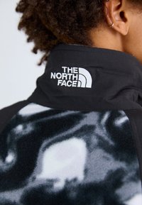 Chaqueta de forro polar negra con un cuello alto, un patrón abstracto en blanco y gris, y un logo blanco destacado en la parte de atrás.