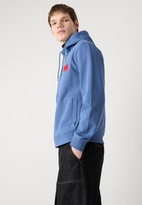 Blå hettegenser laget av en bomullsblanding, med en frontlomme, ribbet mansjetter og en rød logo-patch på brystet. Mørke jeans brukt under.