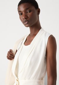 White sleeveless top with a V-neck design, soft fabric, and clean lines. The model holds a light-colored bag, showcasing simple accents.