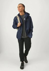 Navy waterproof jacket with hood, zippered front. Dark gray t-shirt underneath. Black pants and sporty shoes. Backpack straps visible.
