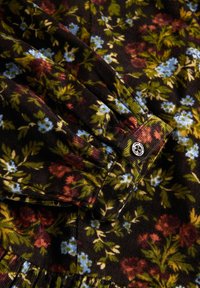 Ciemny materiał z gęstym, kwiatowym wzorem w odcieniach zieleni, niebieskiego i rdzy, pokazujący marszczony mankiet rękawa z okrągłym przyciskiem z logo.