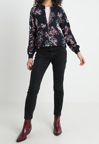 Vrouw in een marineblauwe bloemenjack met rits, zwarte jeans en glanzende bordeauxrode enkellaarzen, staand tegen een effen achtergrond.