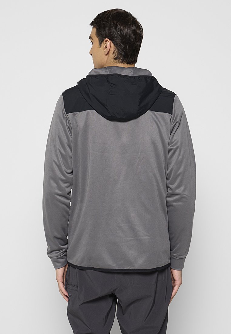 Ein Mann, der sich von hinten zeigt und eine graue Jacke mit langen Ärmeln und einer schwarzen Kapuze sowie dunkle graue Hosen trägt, vor einem schlichten hellen Hintergrund.