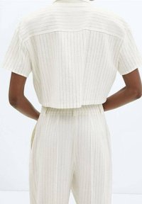 Chemise à manches courtes de couleur crème avec des rayures verticales subtiles, présentant une coupe courte et une fermeture au dos par boutons pour un look ajusté.