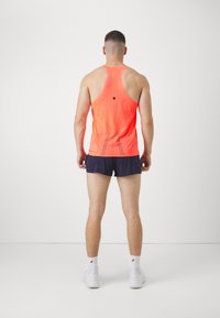Mann in einem leuchtend orangefarbenen, durchlöcherten Tanktop, dunklen Sportshorts, weißen Socken und weißen Turnschuhen, der mit dem Gesicht zu einer weißen Wand steht.