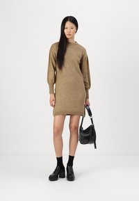 Beige stickad klänning med långa ärmar, ribbat fåll och en avslappnad passform. Kombinerad med svarta platåsandaler och en liten svart axelväska.
