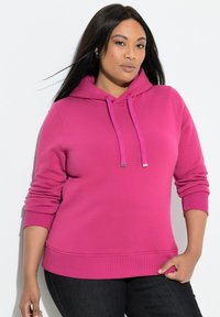 Kobieta ubrana w jaskraworóżową bluzę z kapturem i sznurkami oraz czarne spodnie, stoi z jedną ręką w kieszeni na białym tle.