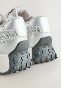 Srebrne buty sportowe z gładką metaliczną cholewką, ozdobione przezroczystymi dżetami, z teksturowaną czarną gumową podeszwą oraz wyściełanym kołnierzem.