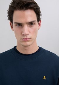Sweatshirt bleu marine foncé à col rond, avec un petit logo jaune de tête de mort et os croisés sur la poitrine gauche. Tissu lisse en mélange de coton.