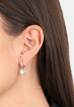 Boucles d'oreilles en argent en forme de croissant, ornées d'un petit pendentif en forme de cœur brillant, mettant en valeur une texture lisse et une finition polie, portées sur le lobe de l'oreille.