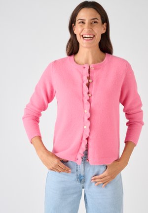 Donna sorridente che indossa un cardigan rosa peloso con bottoni e jeans azzurri, in piedi con le mani vicino alle tasche davanti a uno sfondo neutro.