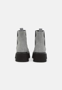 Vaaleanharmaat mokkanilkkurit, joissa mustat sivupaneelit ja paksut kumipohjat. Varustettu vetolenkkeillä helpottamaan pukemista ja koristeltu ompeluilla.