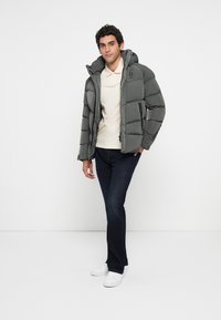 Chaqueta gris acolchada con capucha, que presenta un cierre de cremallera y bolsillos laterales. Lleva puesta sobre una blusa de color claro y se combina con jeans oscuros.