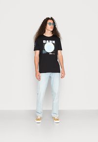 Čierne grafické tričko s okrúhlym svetlo modrým dizajnom a textom, skombinované so svetlo modrými džínsami a hnedými topánkami, na neutrálnom pozadí.