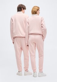 Δύο άτομα στέκονται δίπλα-δίπλα, φορώντας ταιριαστά παστέλ ροζ φούτερ και παντελόνια με λευκά αθλητικά παπούτσια, γυρισμένα με την πλάτη στην κάμερα.