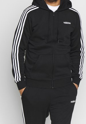 Man draagt een zwarte Adidas trainingspak met witte strepen op de mouwen en broek, handen in de zakken, staande tegen een lichtgrijze achtergrond.