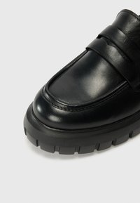 Black leather loafer with a thick rubber sole, featuring a wide strap across the vamp and smooth texture with subtle stitching details.