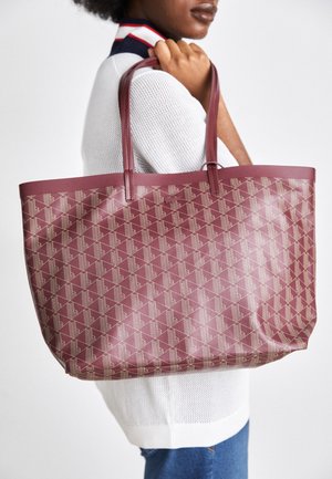 Persoon die een witte gebreide trui draagt, houdt een grote kastanjebruine tas met beige geometrisch patroon over de schouder.