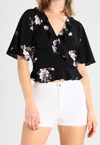 Schwarze Bluse mit floralem Muster und Rüscheneinsatz am Hals und an den Ärmeln, die mit helllila und grünen Blumenmotiven verziert ist, kombiniert mit weißen Shorts.