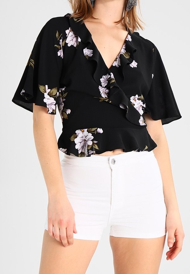 Schwarze Bluse mit floralem Muster und Rüscheneinsatz am Hals und an den Ärmeln, die mit helllila und grünen Blumenmotiven verziert ist, kombiniert mit weißen Shorts.