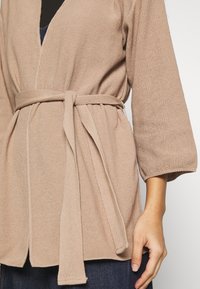 Beige gebreide cardigan met een strikceintuur, driekwartmouwen en een gestructureerde afwerking. De stof lijkt zacht en comfortabel.