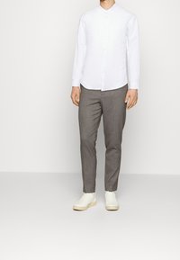 Camicia bianca a maniche lunghe con colletto alla coreana, abbinata a pantaloni grigi chiari su misura e sneakers bianche su uno sfondo neutro.