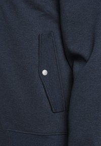 Tissu bleu marine texturé avec un détail de poche visible, un bouton-pression en métal argenté, et une apparence douce et brossée, montrant un gros plan de vêtements.