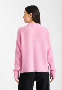 Różowy sweter z dzianiny o ribbowanej fakturze, z wysokim kołnierzem i długimi rękawami. Noszony z czarnymi spodniami, przedstawiający widok z tyłu.
