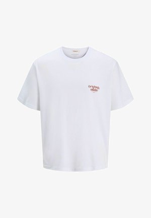 Jongen glimlacht, draagt een wit t-shirt met de tekst "Originals Studio", denim shorts, witte sokken en zwart-witte sneakers, staand op een witte achtergrond.