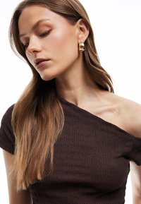 Pendientes en tono dorado con un diseño suave y redondeado. Parte superior texturizada en marrón oscuro con un hombro descubierto, que presenta un patrón acanalado.