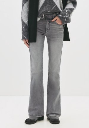 Personne portant un jean flare gris taille haute, un pull gris et blanc à motif, une écharpe noire et des bottes noires, une main dans la poche.