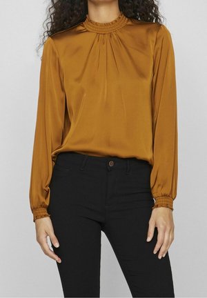 Femme portant un chemisier en satin moutarde à col haut froncé et poignets, associé à un pantalon noir ajusté sur fond uni.