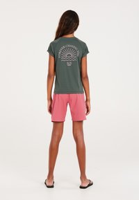 Donkergroene T-shirt met korte mouwen met de tekst "Voor altijd achter de zon aan" en een roze golfontwerp, gecombineerd met roze textuurschoenen en sandalen.