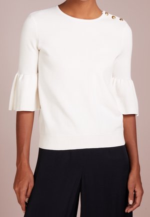 Personne portant un chemisier blanc avec des manches évasées mi-longues au niveau des coudes et des boutons dorés sur l'épaule gauche, associé à un pantalon noir.