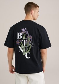 Fekete póló, nagy grafikával, amelyen lila íriszek zöld szárakkal és fehér betűkkel van a "BOTANIC" felirat. Rövid ujjak és laza kialakítás jellemzi.