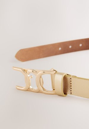 Zelta toņa sprādze ar savstarpēji savienotu dizainu, pievienota šaurai, teksturētai bēšai ādas siksnai. Piecas caurumi izmēra regulēšanai.