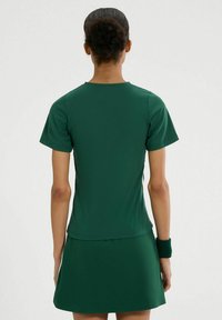 Ciemnozielony top sportowy z krótkim rękawem, wykonany z gładkiego, elastycznego materiału. Prosty design, okrągły dekolt, w zestawie z dopasowaną spódnicą i opaską na nadgarstek.