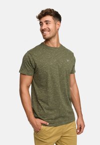 Zielony t-shirt z krótkim rękawem o subtelnej fakturze, okrągłym dekolcie i małym logo na piersi, w połączeniu z jasnym kolorem spodni.