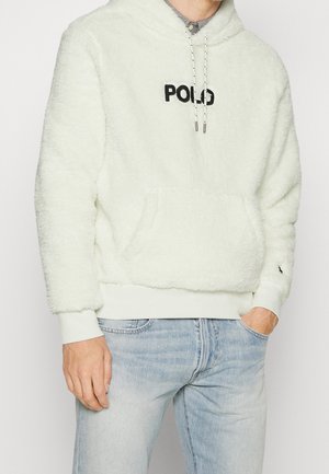 Man draagt een witte, pluizige hoodie met de tekst "POLO" in het zwart, lichtblauwe spijkerbroek en heeft zijn handen deels in de zakken, tegen een effen achtergrond.