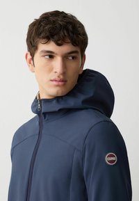 Giacca blu navy con colletto alto e cappuccio, caratterizzata da una zip frontale e una patch con logo circolare sulla spalla sinistra. Tessuto liscio e resistente.