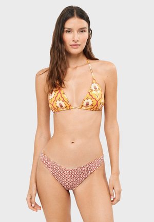 Kobieta w żółtym kwiecistym bikini z wiązaniem na szyję i czerwonych wzorzystych majtkach bikini, stojąca na jednolitym jasnoszarym tle.