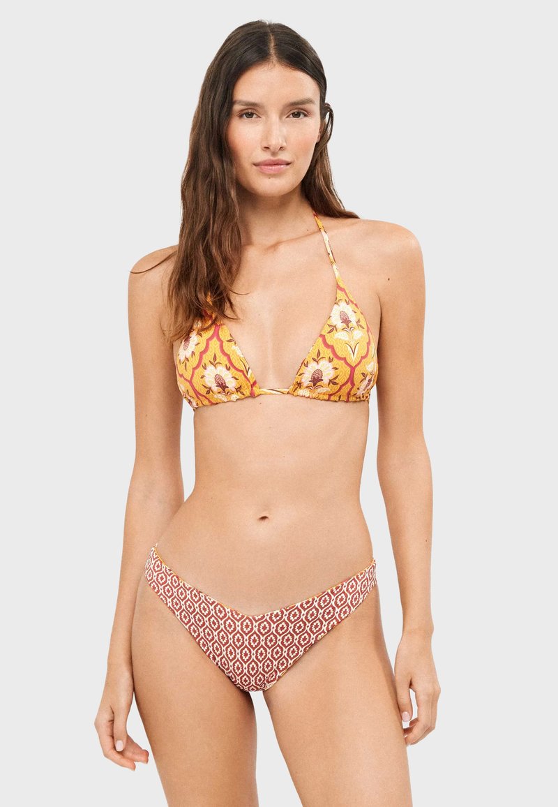Femme portant un haut de bikini jaune à fleurs style dos-nu et un bas de bikini rouge à motifs, debout devant un fond uni gris clair.