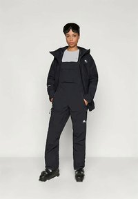 Schwarze isolierte Skijacke mit Kapuze, kombiniert mit schwarzen Latzhosen. Verfügt über Logo-Akzente, Taschen und Skibrille. Unifarbiges Design.