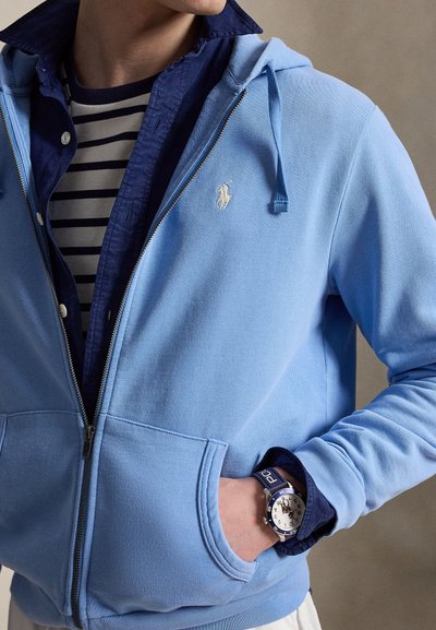 Vīrietis valkā gaiši zilu kapuci džemperi ar rāvējslēdzēju virs tumši zilas pogātas krekls un svītrains T-krekls, uz kreisās rokas plaukstas pulksteni un roku kabatā.