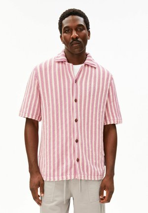 Mann mit kurzen lockigen Haaren und Schnurrbart, der ein rosa-weiß gestreiftes, kurzes Strickhemd mit Knöpfen und hellgraue Hosen trägt.