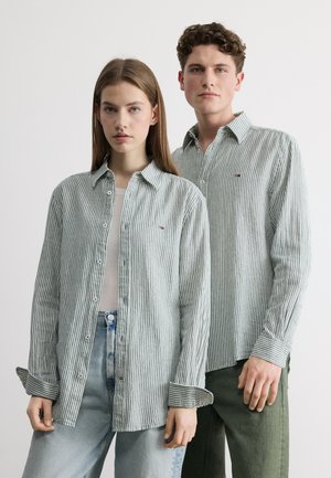 Młoda kobieta i mężczyzna ubrani w pasiaste na pionowo koszule na guziki, stojący obok siebie na jednolitym tle.