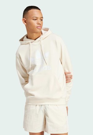 Beige hoodie met een voorkantvak en wit logo. Lichtgewicht stof, capuchon met trekkoord, gecombineerd met lichte shorts. Soepele textuur.