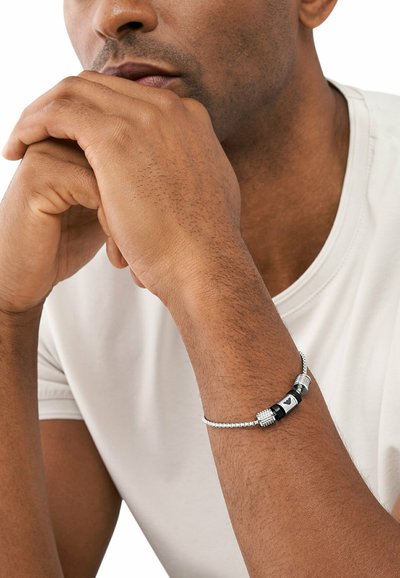Silberfarbener Armreif mit strukturierten Metallakzenten und einer schwarzen zentralen Perle mit einem Muster. Am Handgelenk getragen mit einem neutralen Hemd.