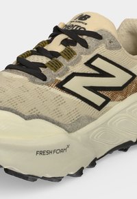 Μπεζ trail παπούτσι New Balance με μαύρα κορδόνια, μεγάλο λογότυπο "N", σόλα Fresh Foam και διακοσμητικές τρύπες κορδονιών σε λευκό φόντο.